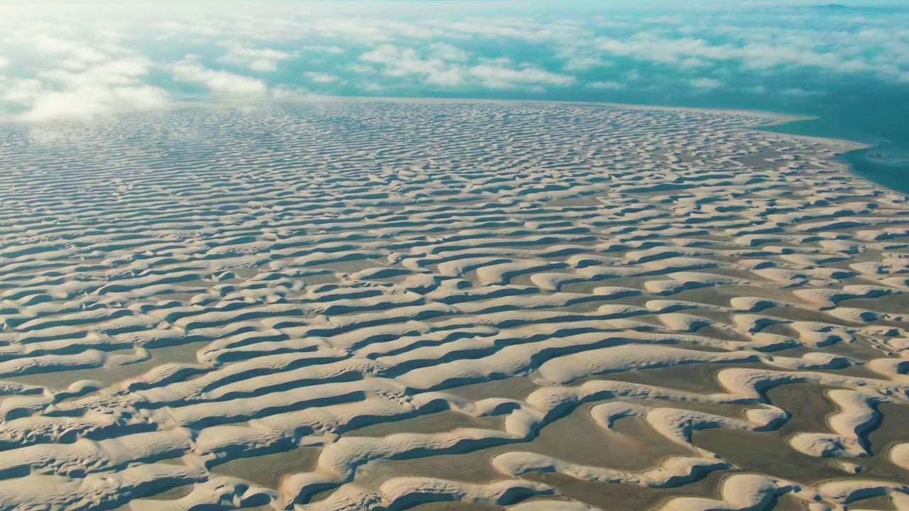 At 0:02, the Guerrero Negro dune field remains dominant, with alternating sunlit sand and shadowed troughs leading toward a faint blue water edge at frame right and no visible people.