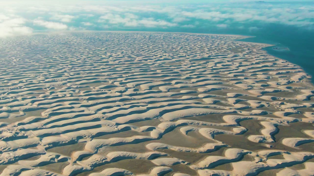 Opening aerial frame over patterned white and tan dunes near the Guerrero Negro coastline, with blue ocean at right, pale clouds above, and no visible people.