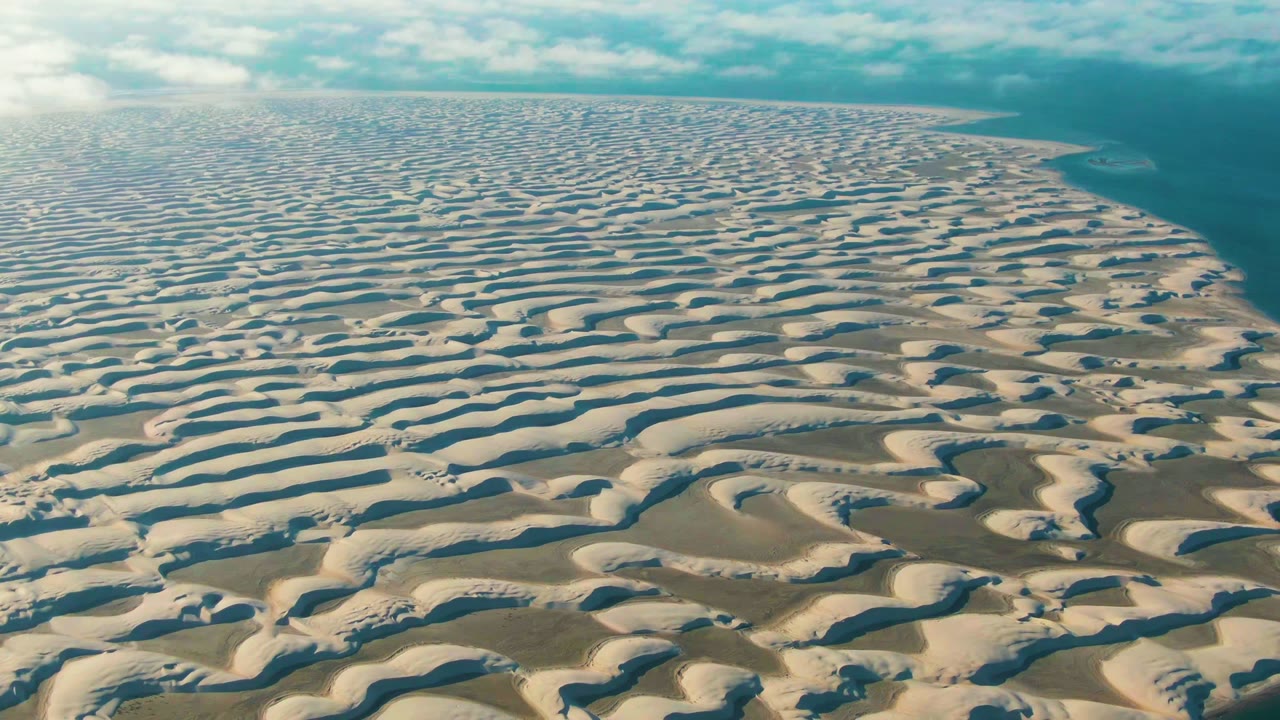Wide aerial frame of Guerrero Negro dunes with rippled white ridges, darker sandy channels, a blue shoreline along the right side, soft cloud cover, and no visible people.