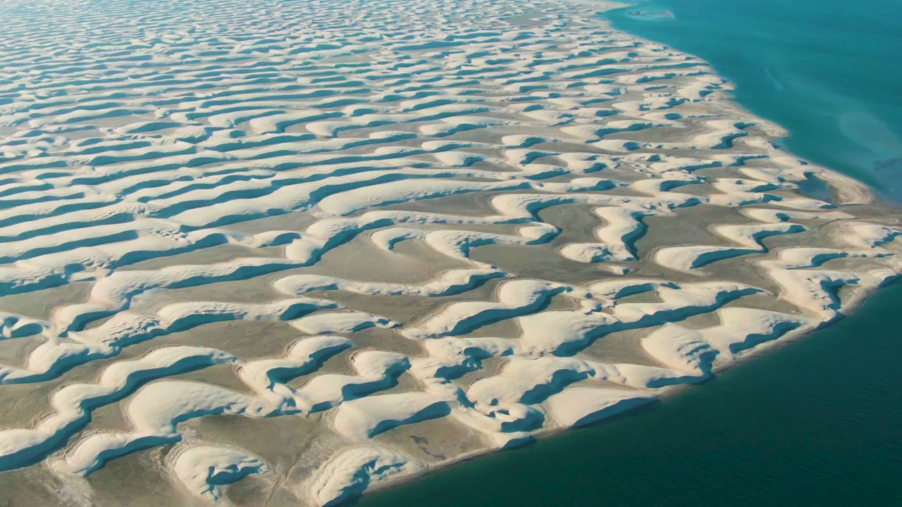 High oblique frame over Guerrero Negro dunes where pale sand waves fill most of the view and dark blue water borders the right side, with no visible people.