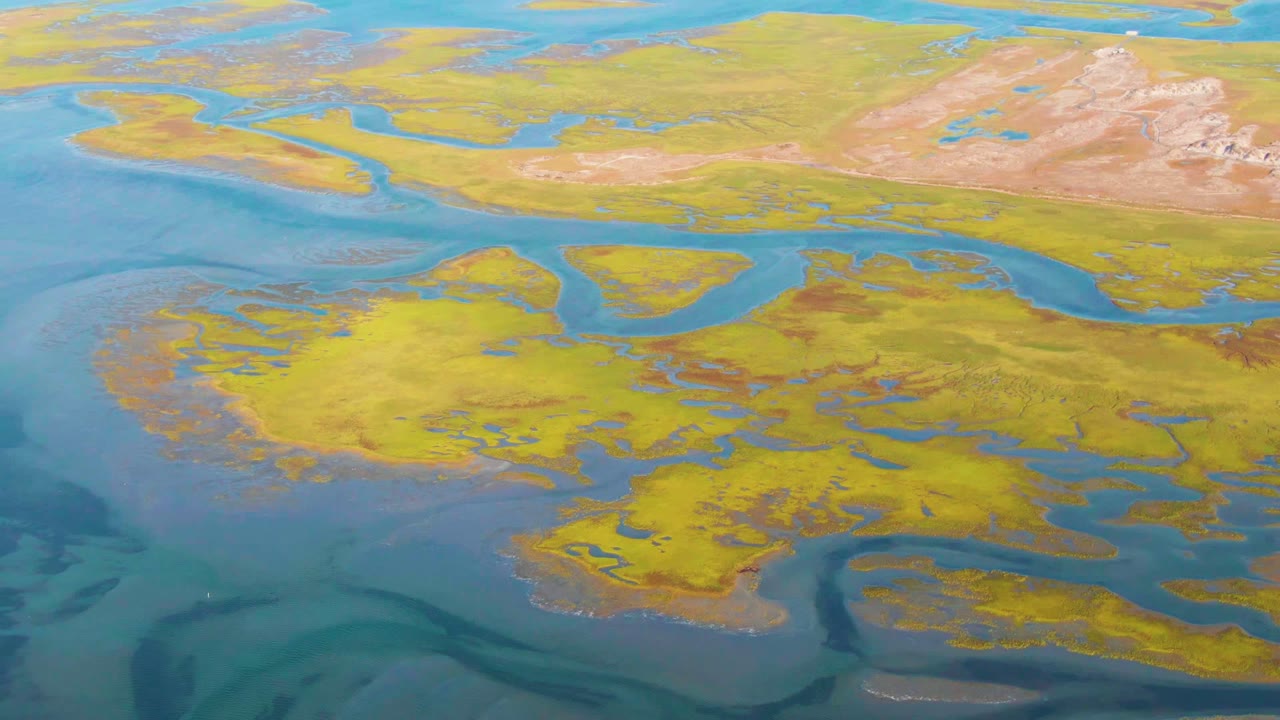 Drone-style aerial view over Guerrero Negro with turquoise water channels, ochre wetland ground, distant pale hills, and no visible people.