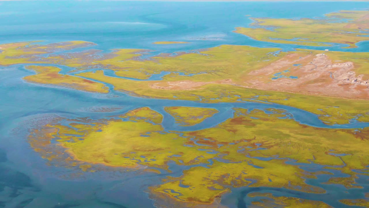 Aerial view in Guerrero Negro with yellow marsh flats, calm blue lagoon water, and pale dry ridges on the right, with no visible people.