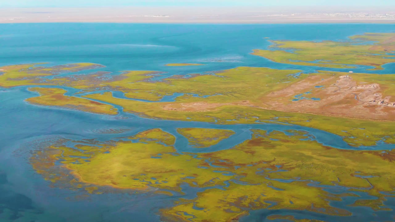 A wide aerial frame over Guerrero Negro coastal wetlands shows blue water, greenish-yellow marsh vegetation, brown ground patches, a pale horizon, and no visible people.