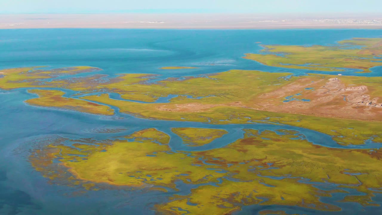 At two seconds, the aerial view holds on Guerrero Negro wetlands where yellow-green vegetation meets open blue water under a light sky, with no visible people.