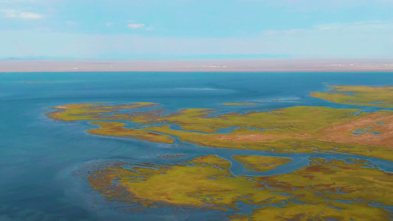 The Guerrero Negro wetland remains in a wide aerial composition, with marsh islands across the middle, darker blue water at left, pale cloud patches, and no visible people.