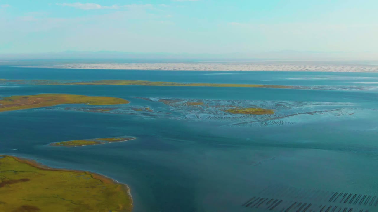 Opening aerial frame over Guerrero Negro coastal wetlands with green marsh land in the lower left, shallow blue-green water, distant flats, and no visible people.
