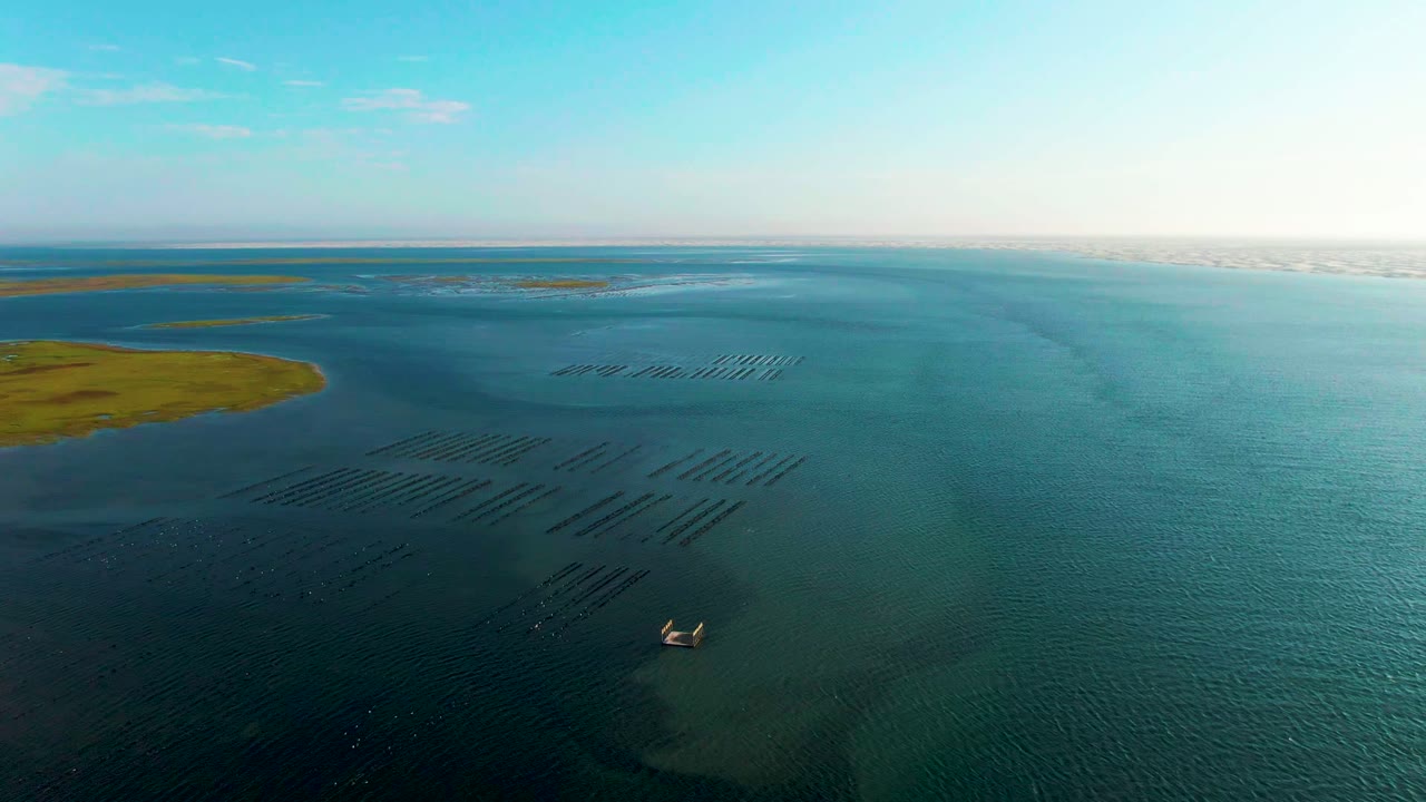 Opening aerial frame over Guerrero Negro, Baja California Sur, showing aquaculture rows in shallow blue-green water, green land to the left, a small boat near the lower center, and no visible people.