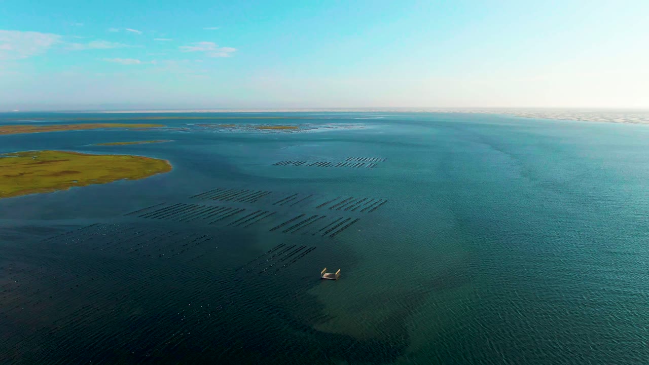 The aerial composition over Guerrero Negro shows aquaculture structures spread across the midground, a larger green landmass entering from the left, the boat low in frame, and no visible people.