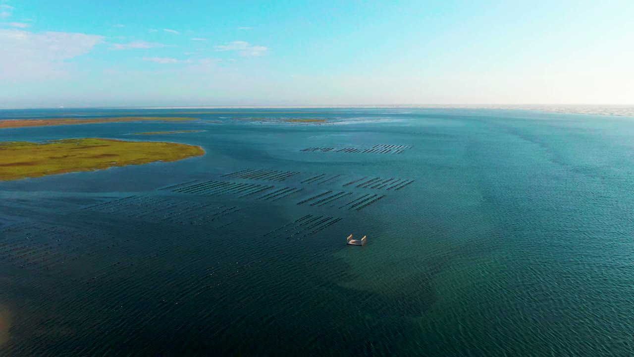 Aerial view over Guerrero Negro, Baja California Sur, with blue-green water, parallel aquaculture rows, two small boats near the lower area, green land at left, and no visible people.