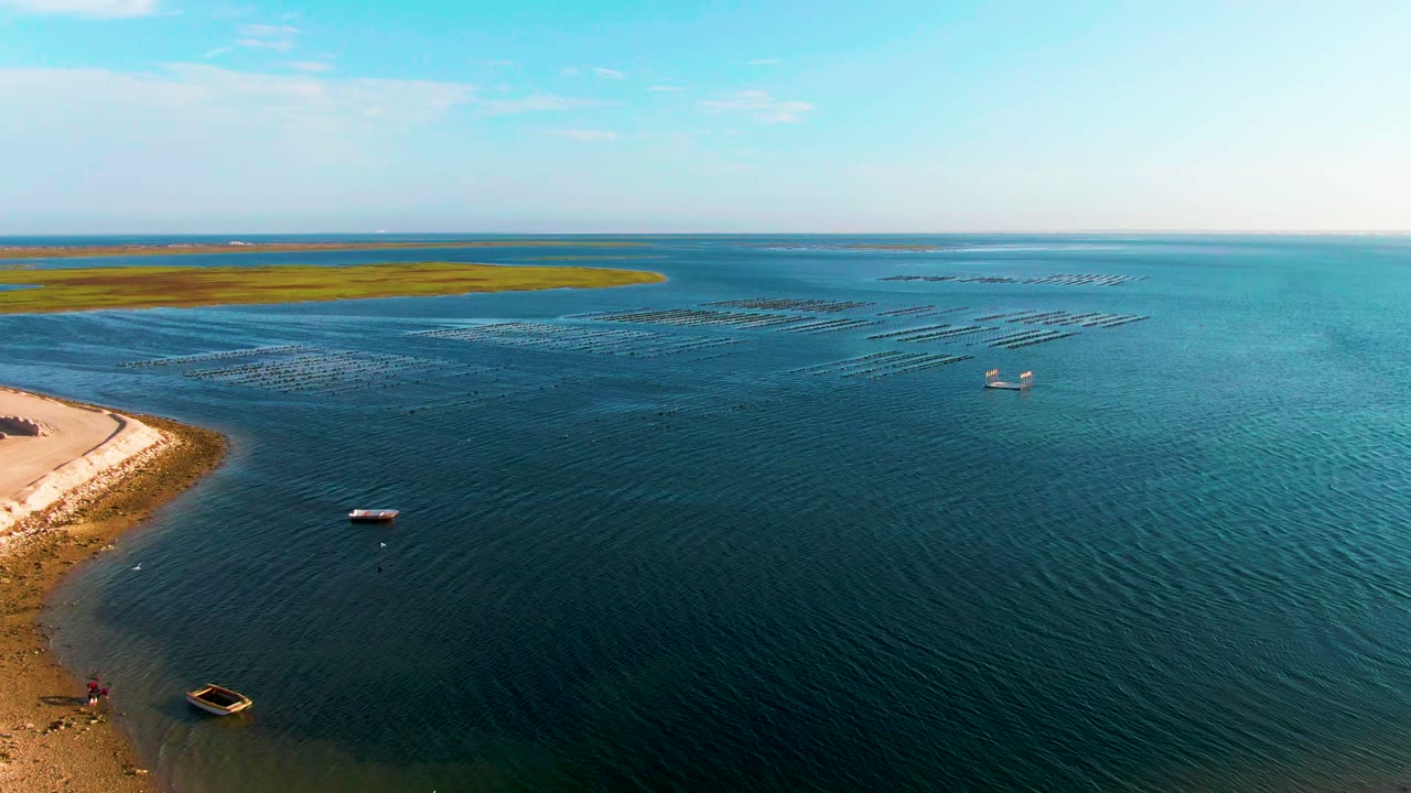Aerial view over Guerrero Negro coastal water, with sandy shore at left, a small boat near the shallows, floating rows offshore, and no visible people.