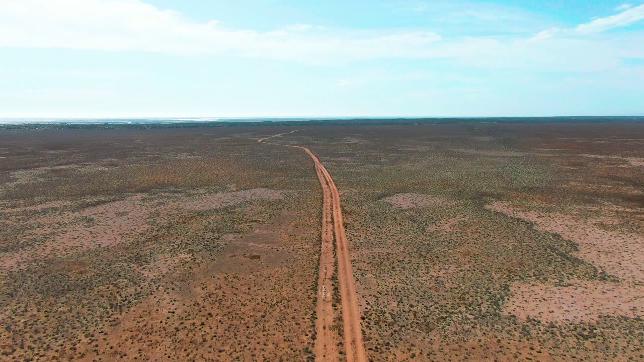Aerial view in Guerrero Negro, Baja California Sur, with a reddish dirt road rising from the lower frame through arid scrubland toward a flat horizon, no visible people.