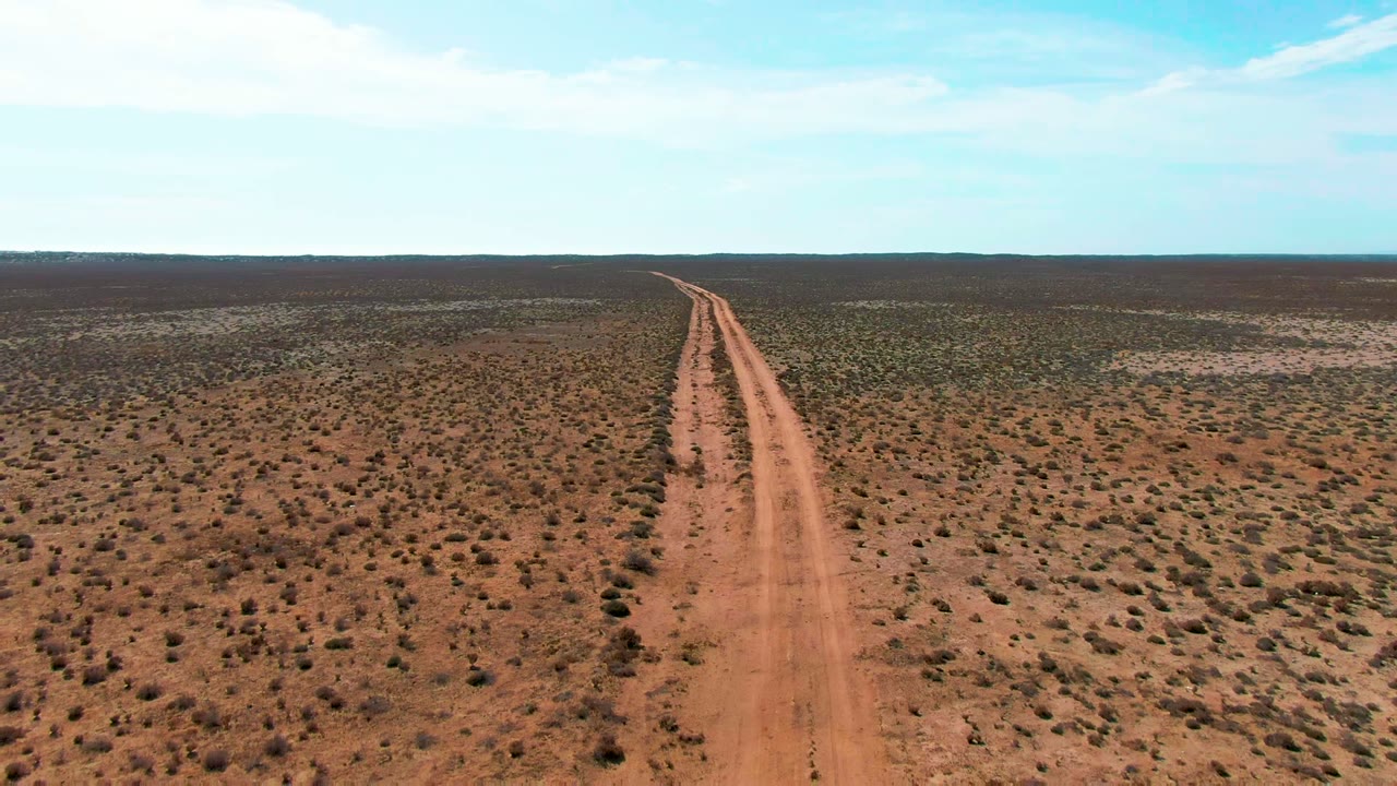 At 0:00, an aerial view in Guerrero Negro, Baja California Sur shows a reddish dirt road starting near the lower center and running toward a flat horizon, with no visible people.