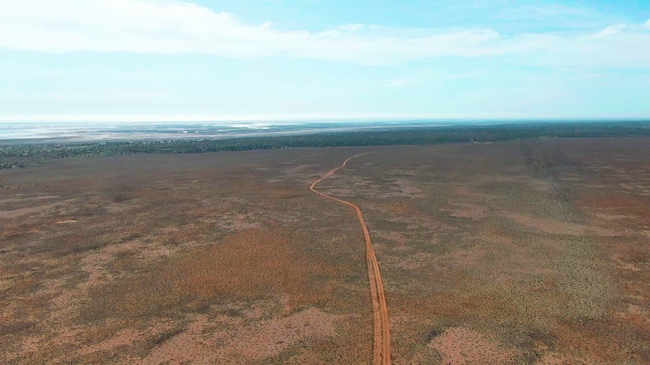 Aerial frame from Guerrero Negro, Baja California Sur showing a reddish dirt road entering from the lower center and curving across dry scrubland under a pale cloudy sky, with no visible people.