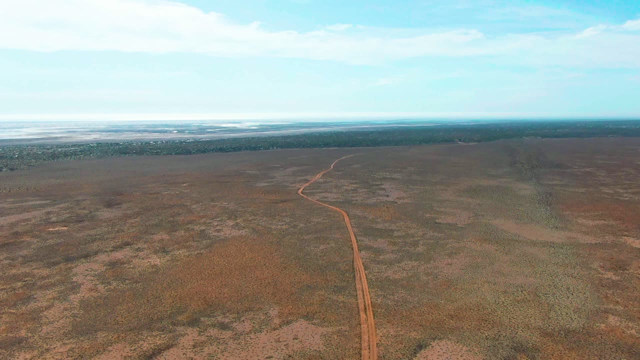 Aerial frame from Guerrero Negro, Baja California Sur showing the reddish road as a narrow vertical path through flat scrubland, with a broad bright horizon and no visible people.