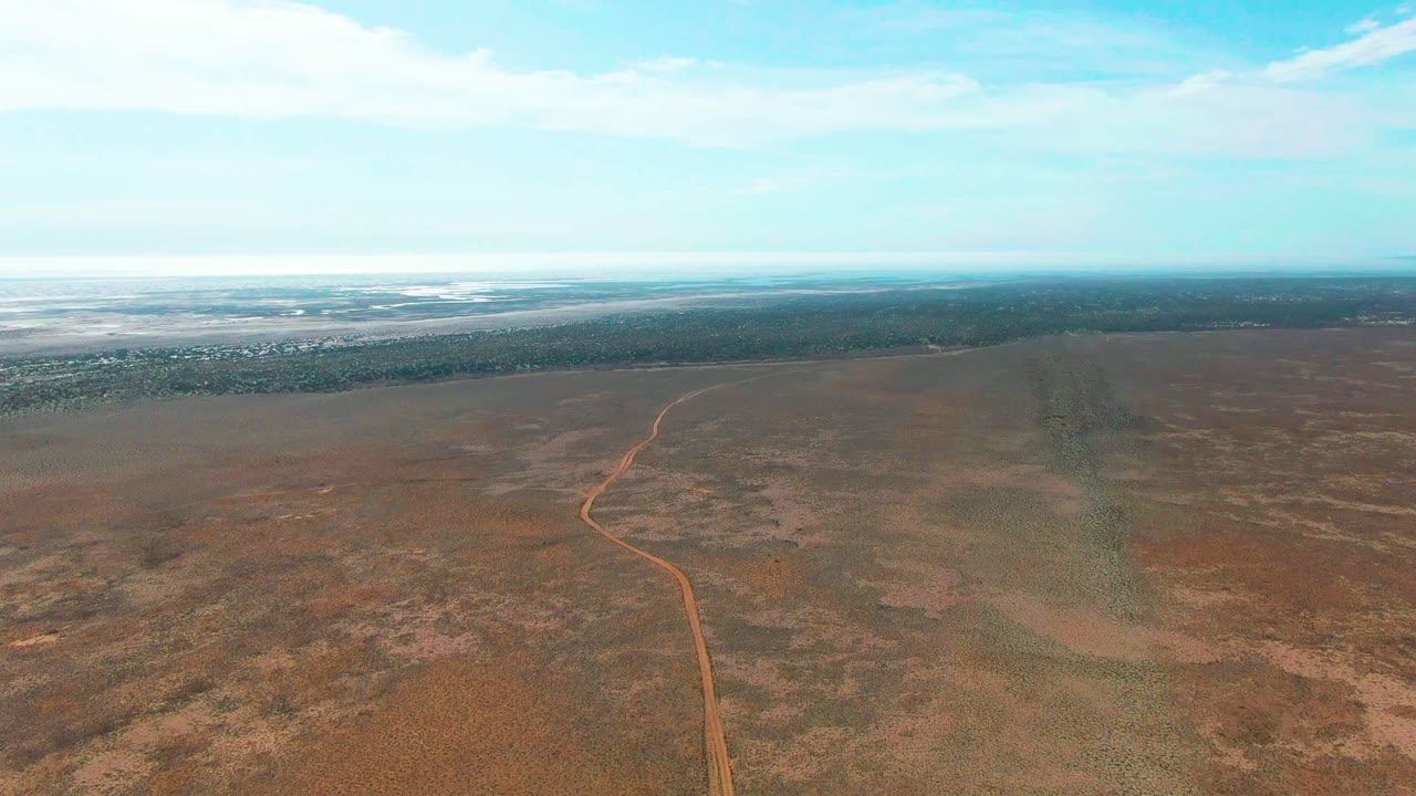 Opening aerial frame in Guerrero Negro, Baja California Sur, with an orange dirt road entering from the lower left and bending through sparse brown scrubland, no visible people.