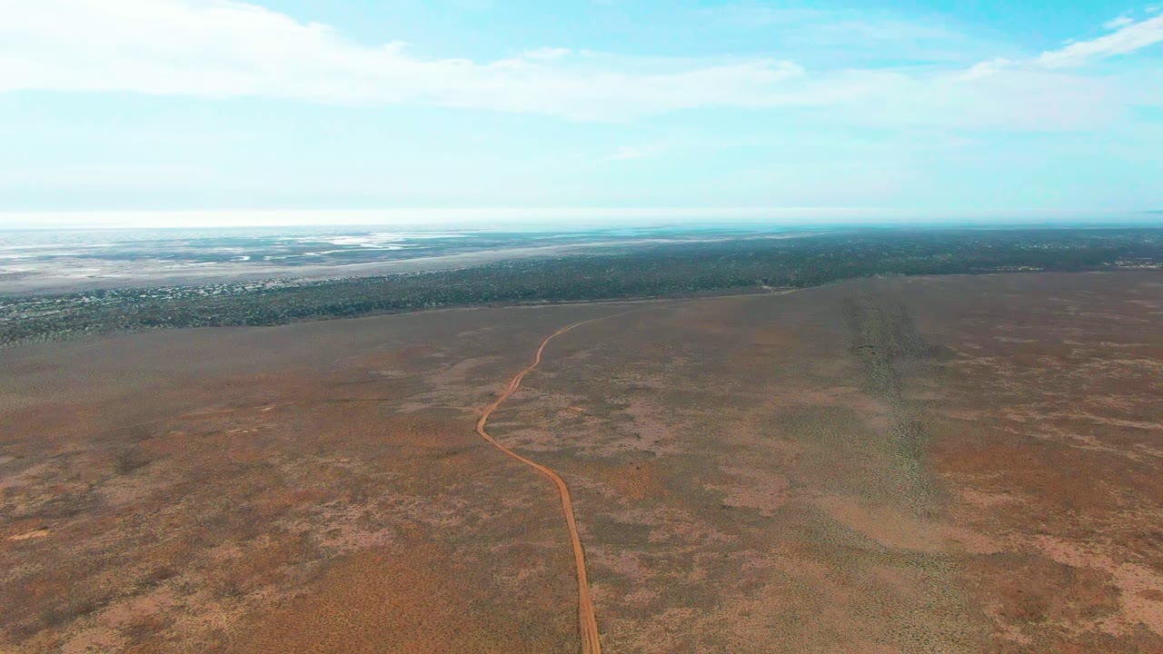 High aerial view of a narrow dirt road crossing dry open land with distant flats.