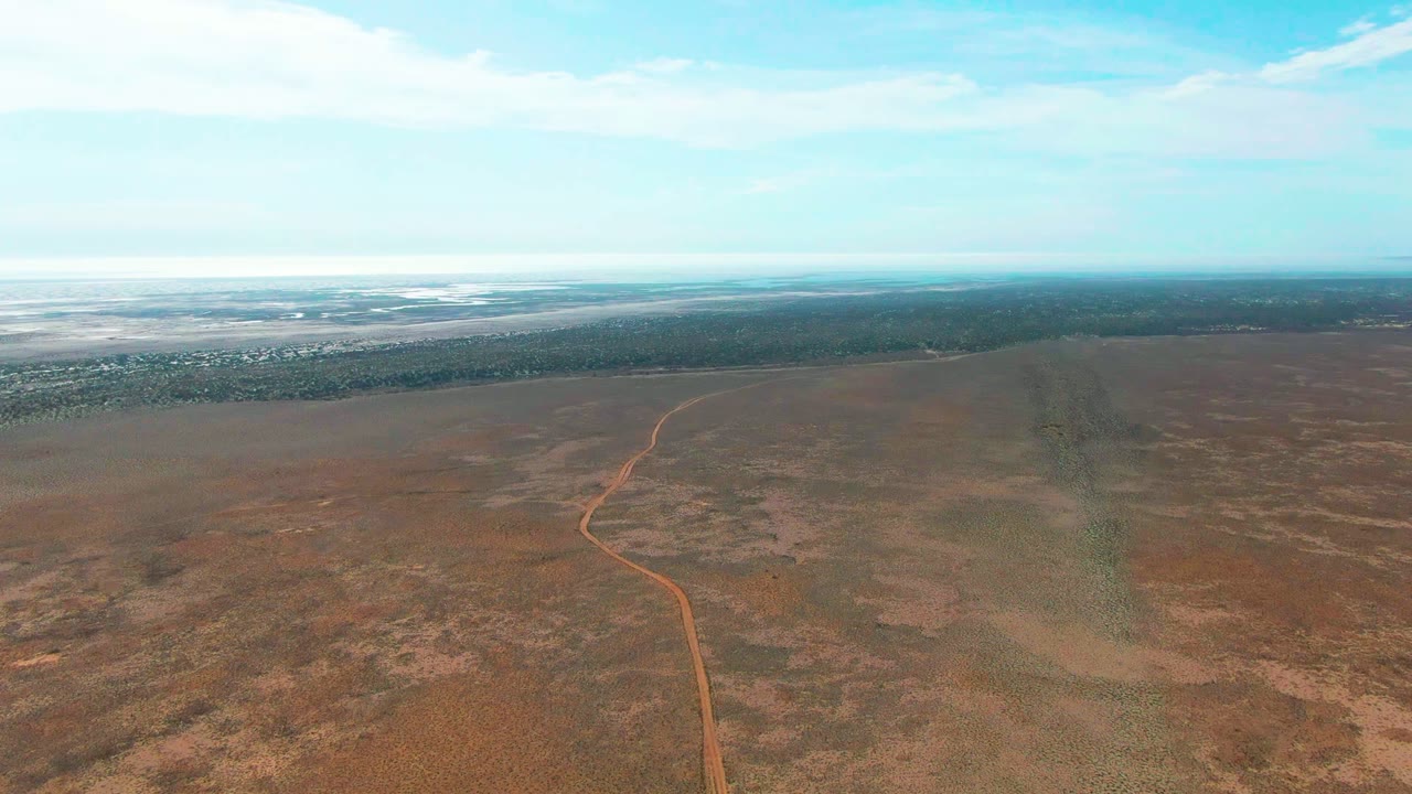 Mid-clip aerial frame in Guerrero Negro, Baja California Sur, with the dirt track curving upward through flat brown vegetation and a broad blue sky above, no visible people.