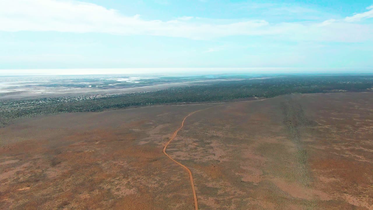 Aerial frame from Guerrero Negro, Baja California Sur, with the reddish dirt track bending upward through open arid terrain, a dark green-brown strip near the horizon, and no visible people.