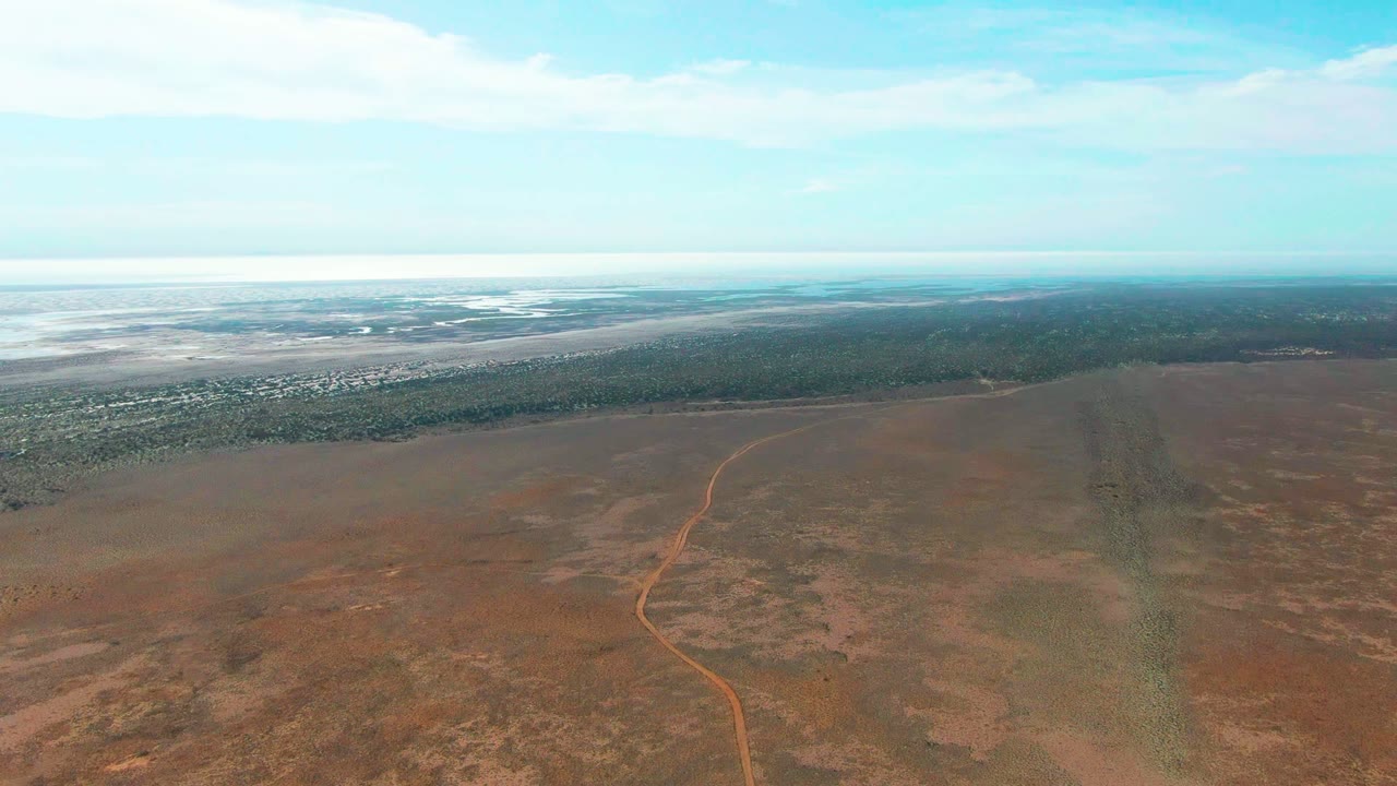 High aerial view of dry open land with an orange dirt track leading toward trees and distant water.