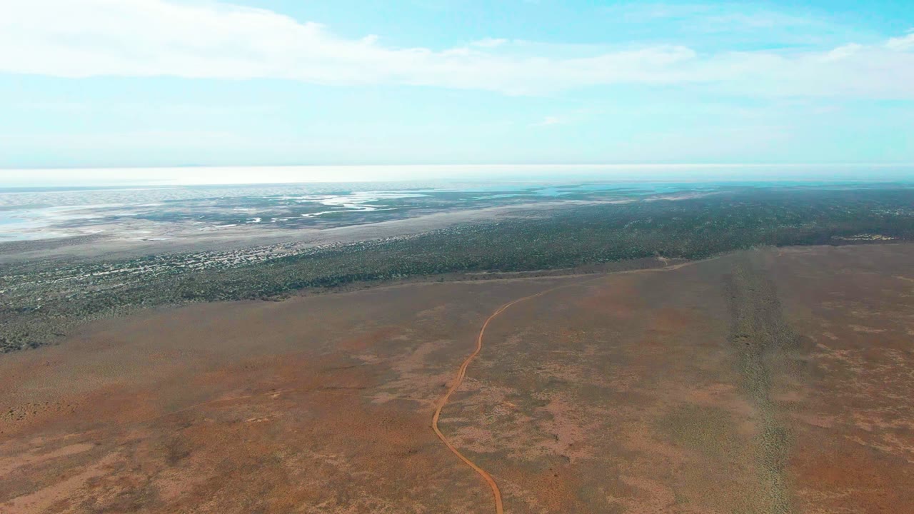 High wide view of a winding dirt road and flat scrubland under blue sky.