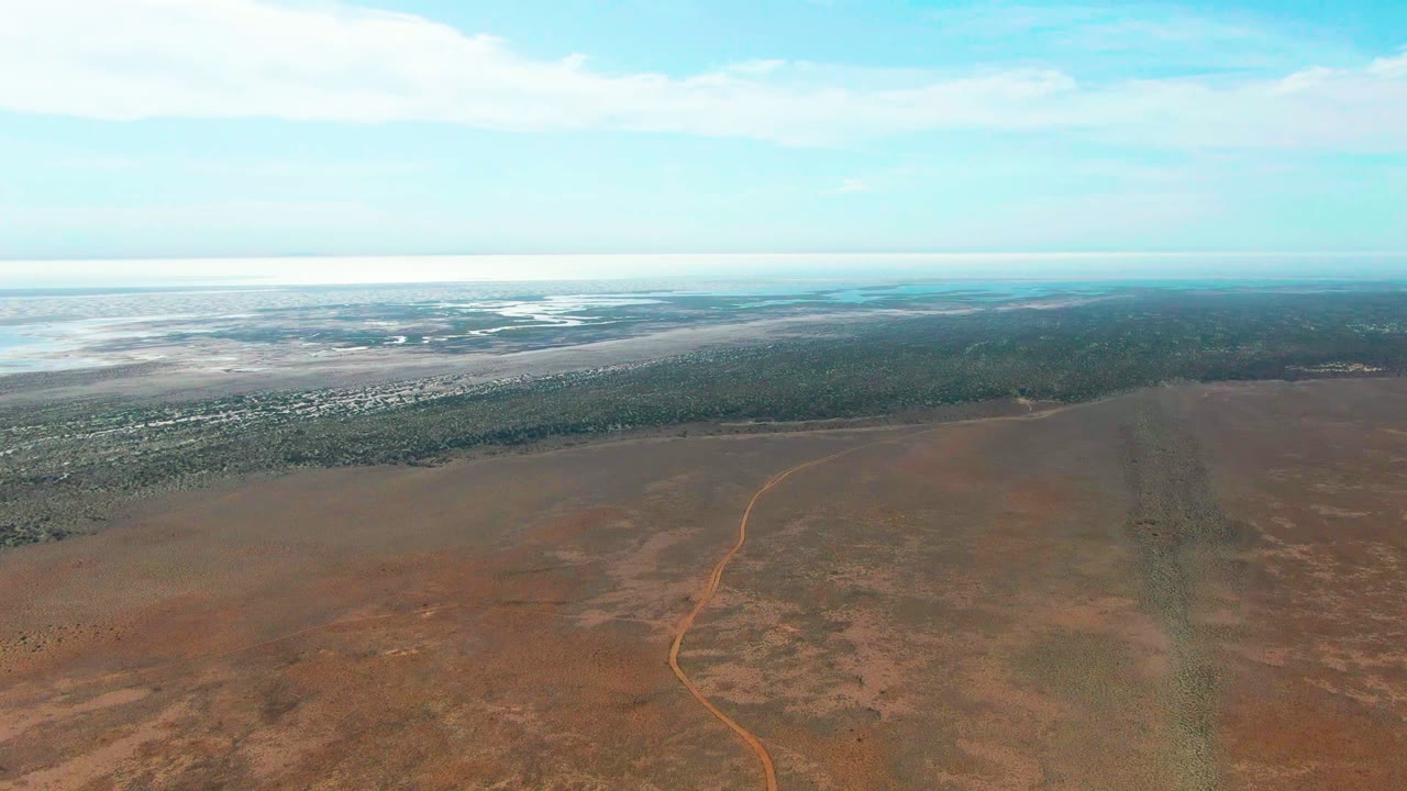 The 0:02 frame holds a broad Guerrero Negro aerial composition with the orange road bending through dry brown ground, dark vegetation across the middle distance, pale water beyond, and no visible people.