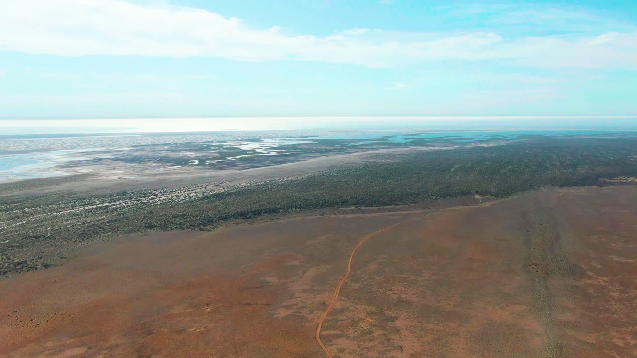 Opening aerial frame over Guerrero Negro, Baja California Sur, with red dry land below, a faint dirt road, green shoreline vegetation, shallow water near the horizon, and no visible people.