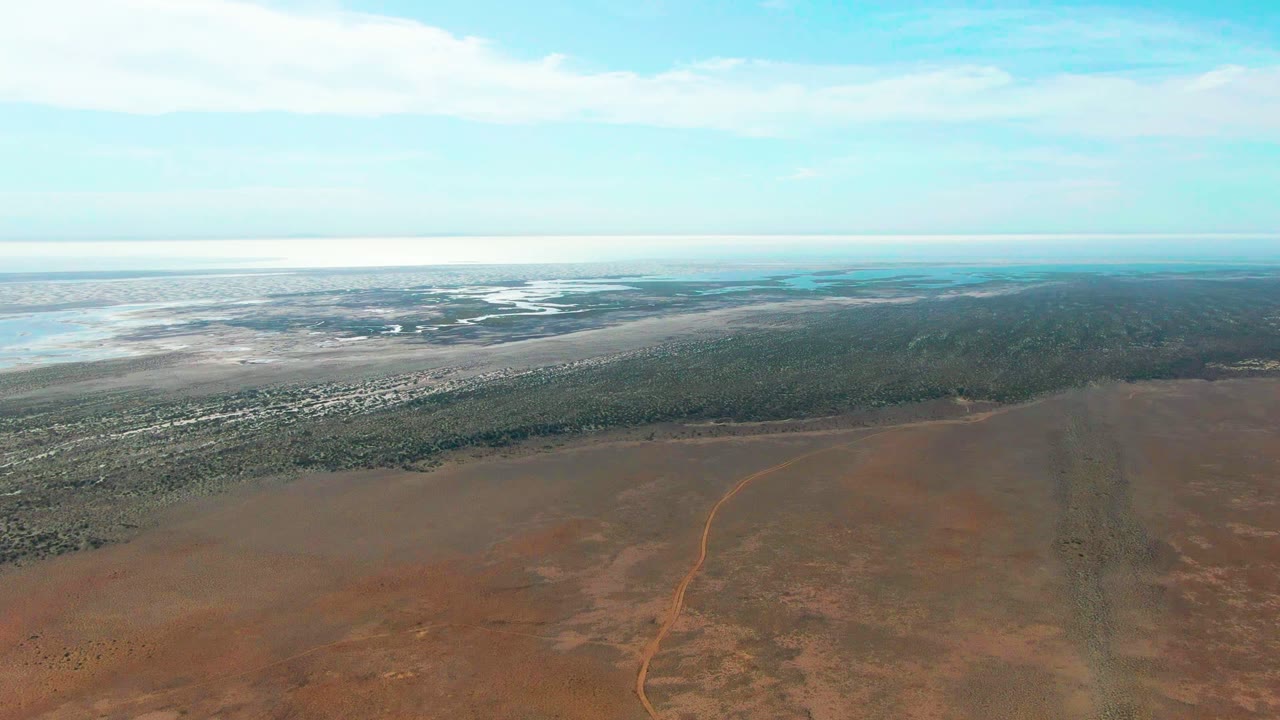 Third frame over Guerrero Negro with red earth filling the lower view, a curving dirt road, a dark green shoreline strip, pale water patches, and no visible people.