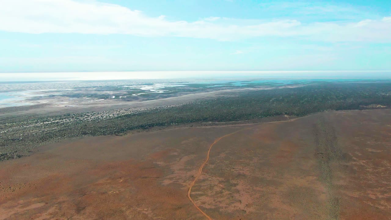 Opening aerial frame over Guerrero Negro, Baja California Sur, showing a red earth plain with a faint dirt road, a dark vegetation band, pale distant water, blue sky, and no visible people.
