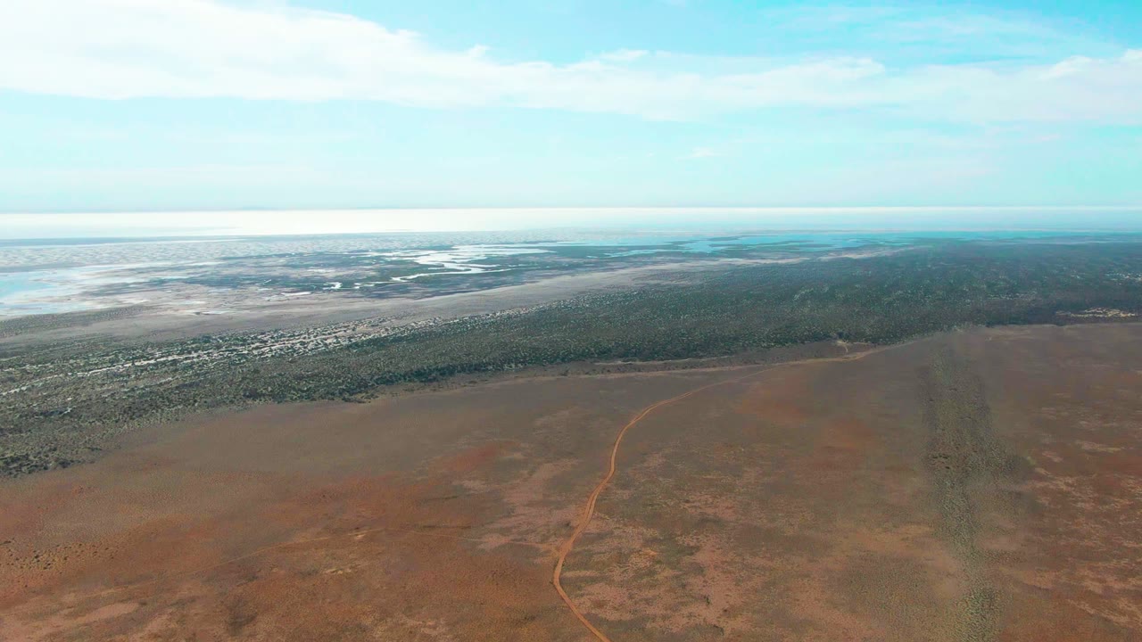 Third frame from the Guerrero Negro aerial view, with the dirt road faintly visible across the red foreground, a broad strip of low green vegetation, distant water, and no visible people.