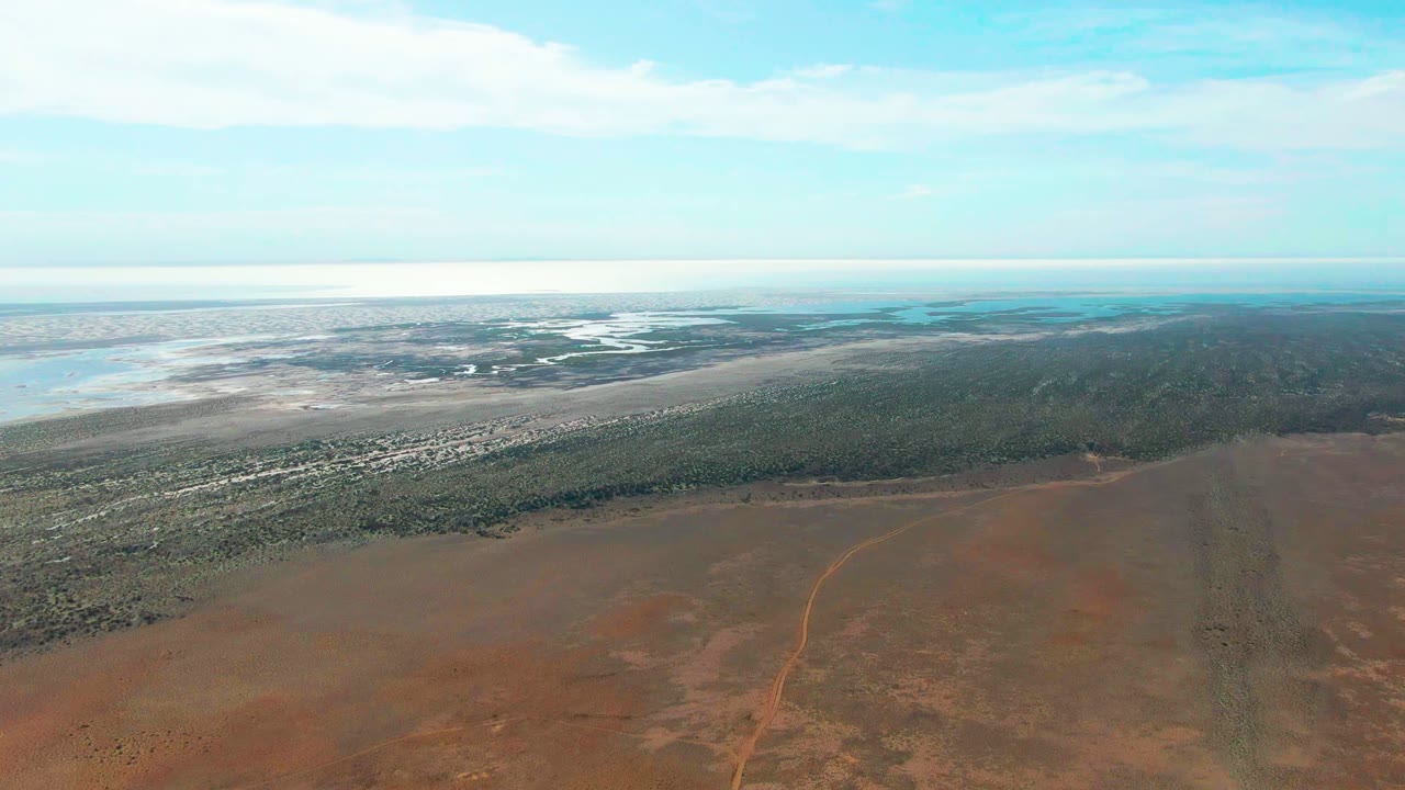 Opening aerial frame over Guerrero Negro, Baja California Sur, with reddish dry ground in front, a green coastal vegetation band, pale wetland water beyond, and no visible people.
