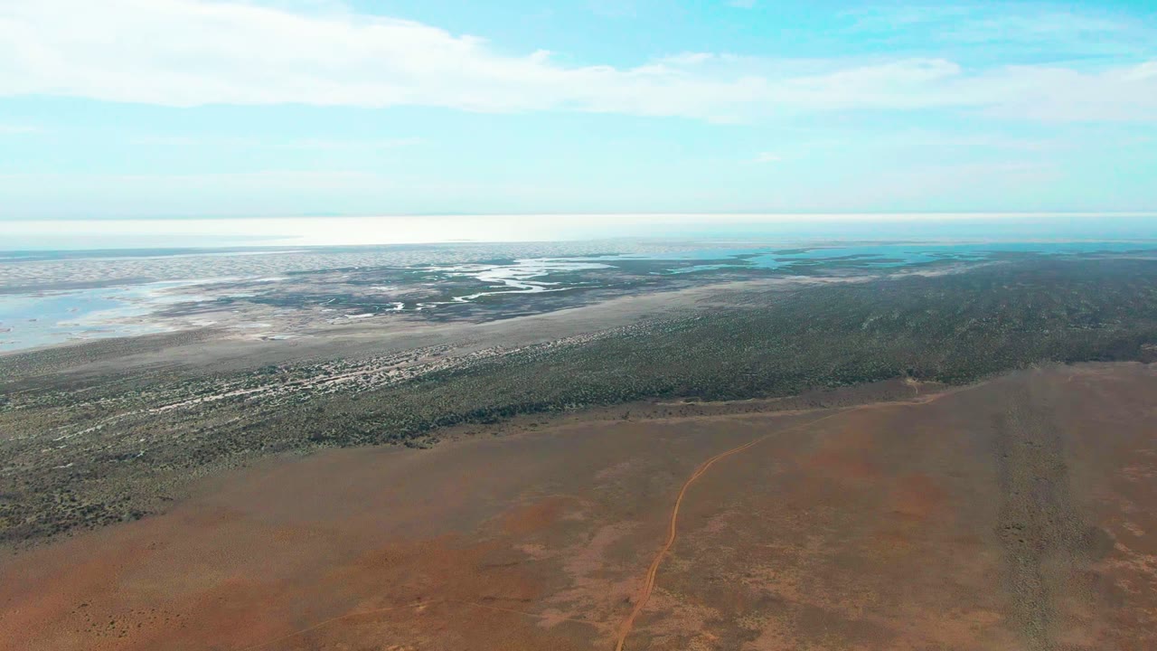 At 0:02, the high view over Guerrero Negro shows a broad reddish foreground, dark green forest across the middle, light-toned water in the distance, and no visible people.