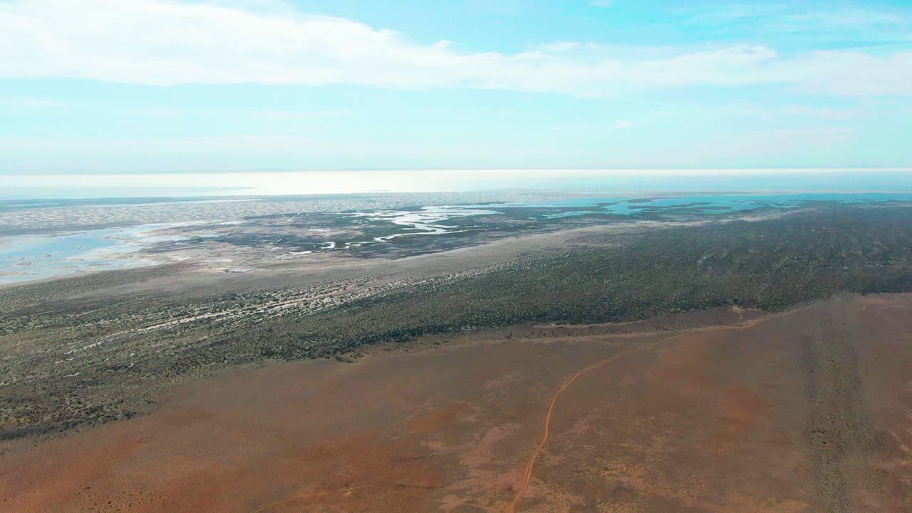 High aerial view over Guerrero Negro showing red-brown desert in the foreground, a green vegetation strip across the middle, pale shoreline flats, blue water, partly cloudy sky, and no visible people.