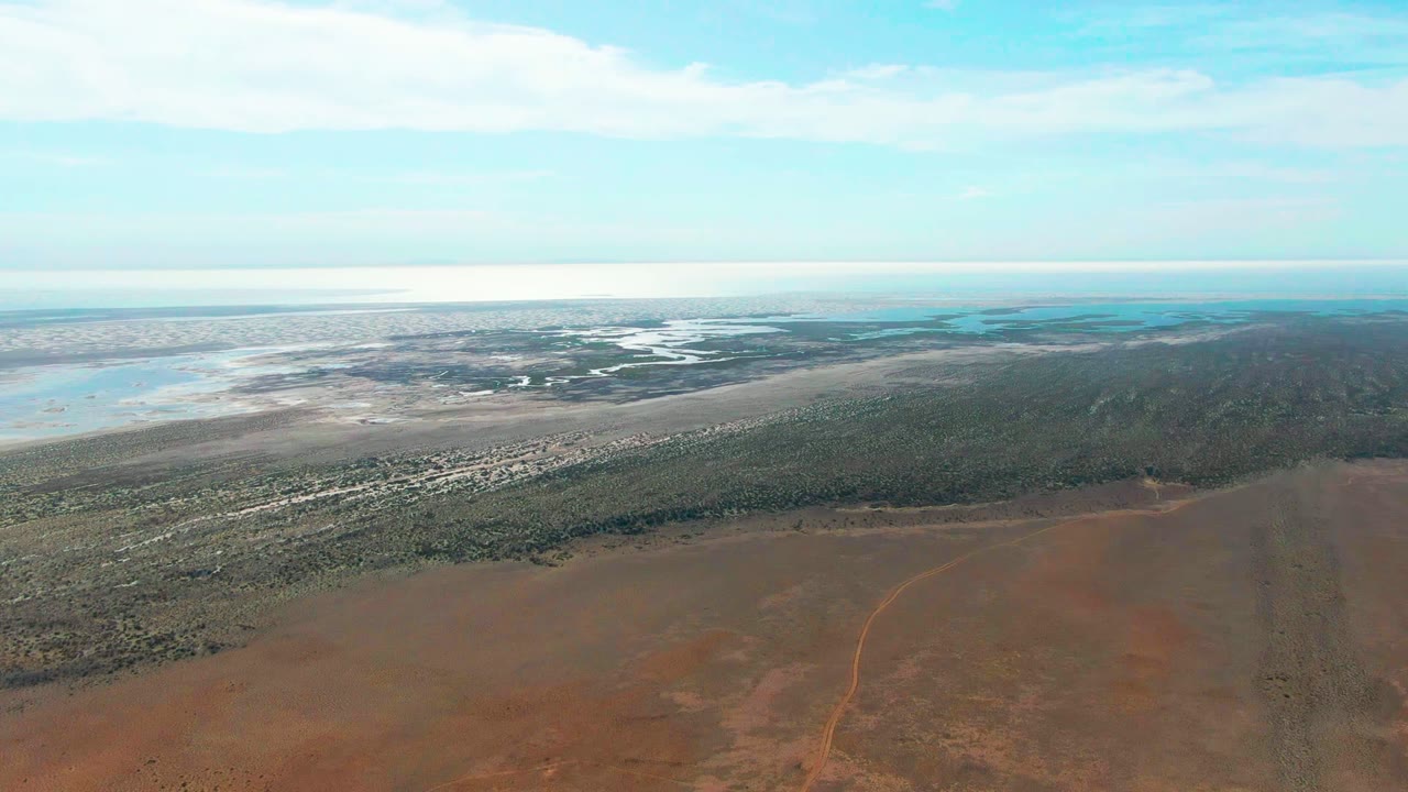 Wide Guerrero Negro aerial looking across dry red ground toward a long green strip, light-colored shoreline flats, blue water on the horizon, thin clouds, and no visible people.