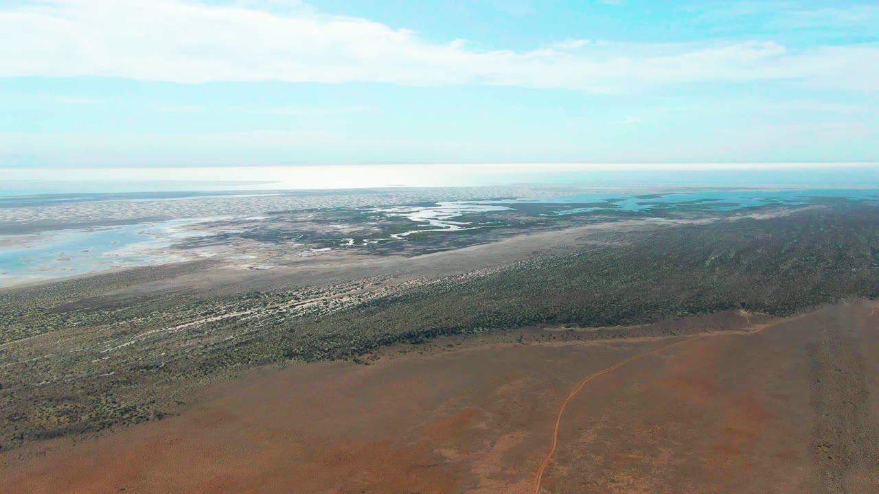 Opening aerial frame over Guerrero Negro, Mexico, with reddish dry terrain below, a green vegetation band, pale wetland water near the horizon, scattered clouds, and no visible people.