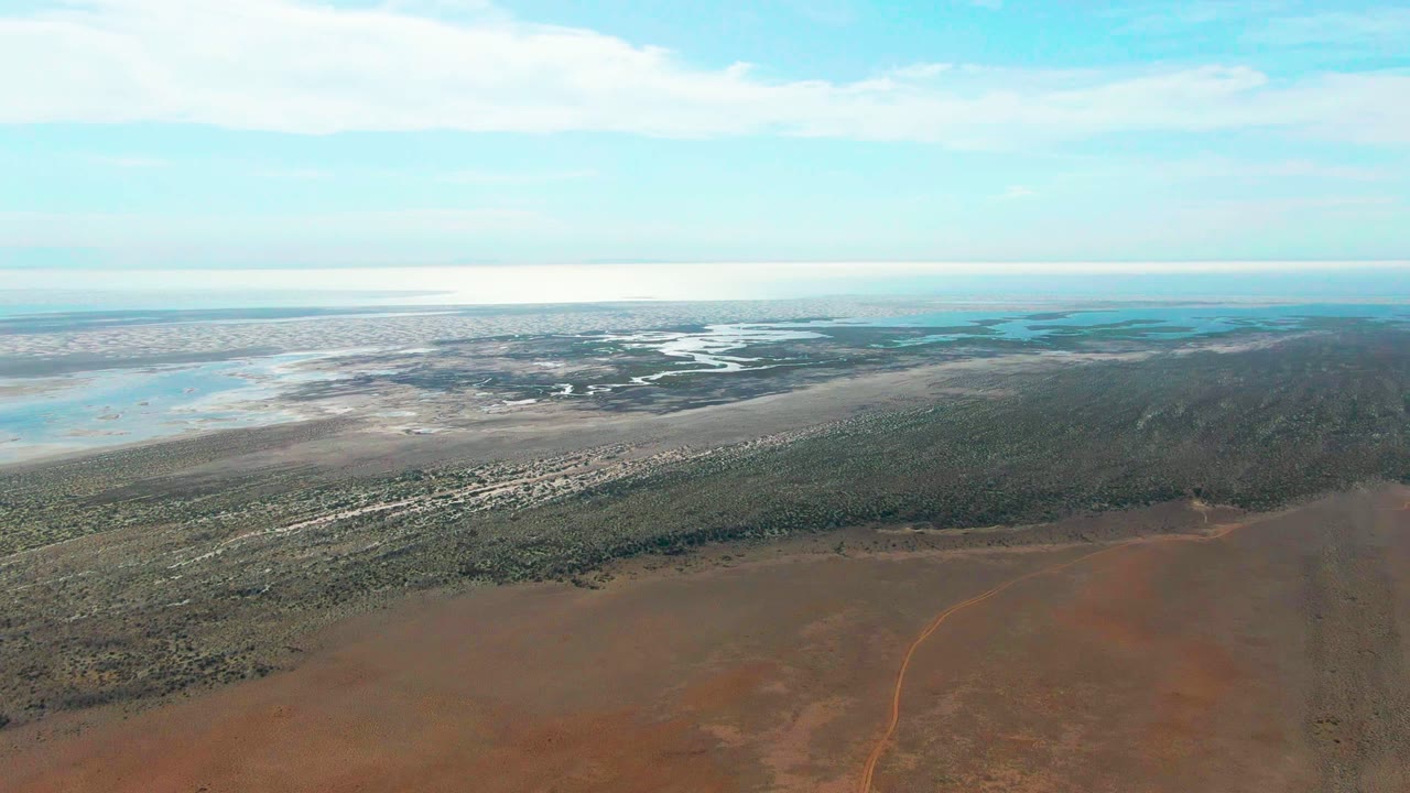 Wide aerial panorama of desert-colored ground, scrub vegetation, wetlands, and cloudy sky.