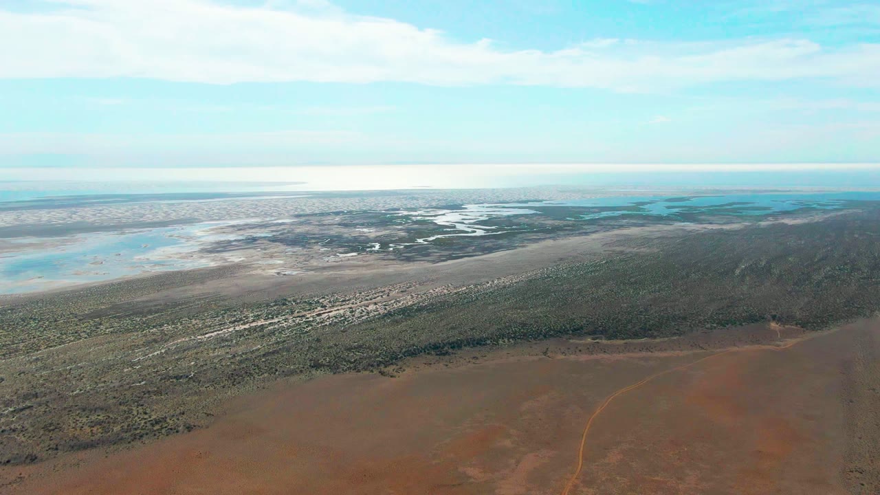 Aerial view over Guerrero Negro, Baja California Sur, with red-brown dry ground in front, green vegetation across the middle, pale wetlands and blue water beyond, and no visible people.