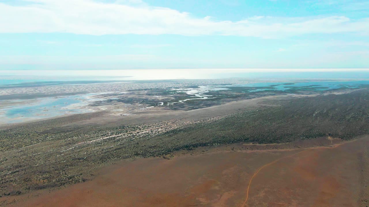 Drone-height view over Guerrero Negro wetlands with red earth at the bottom of frame, green vegetation through the center, pale reflective water flats in the distance, and no visible people.