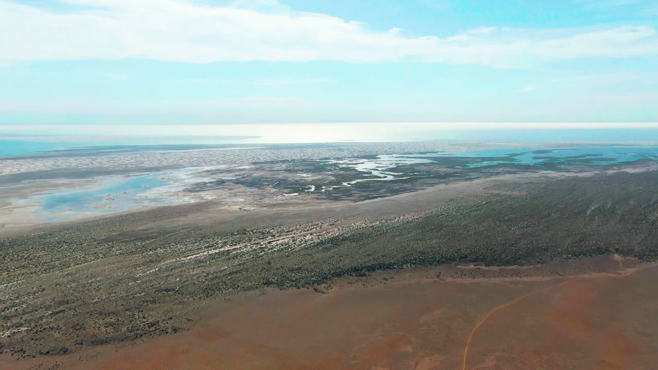 Opening aerial frame over Guerrero Negro, Baja California Sur, with reddish dry ground in front, green vegetation across the middle, pale wetland water in the distance, and no visible people.