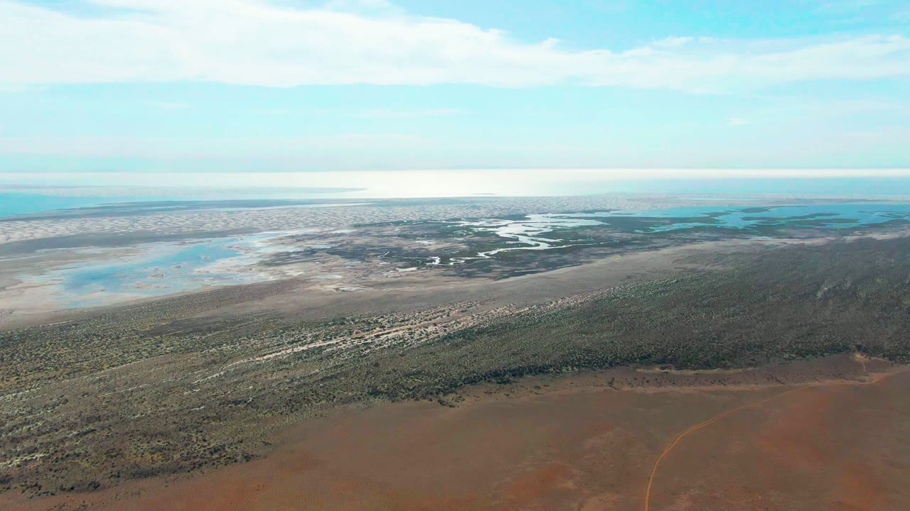 Wide drone view of scrubland leading to pale water and marsh under scattered clouds.