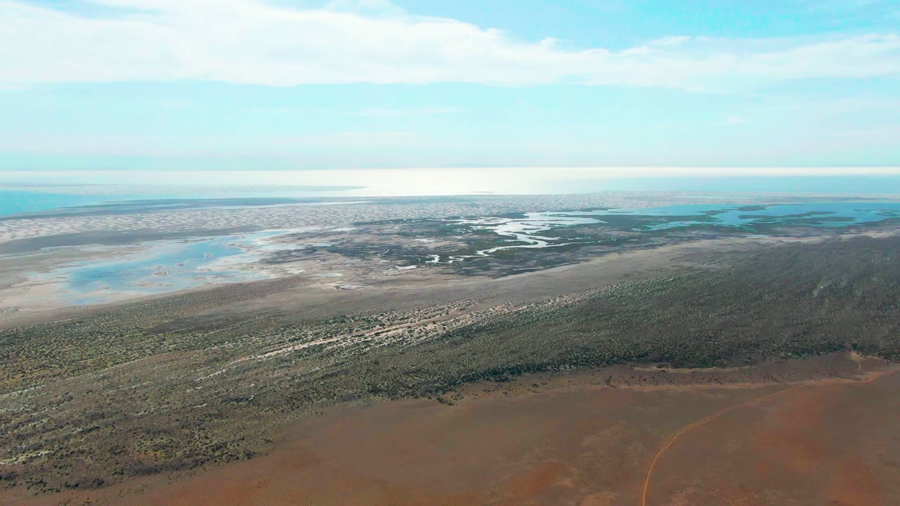 Mid-clip aerial frame over Guerrero Negro with the brown foreground road trace or track faintly visible, green vegetation through the center, pale water behind it, and no visible people.