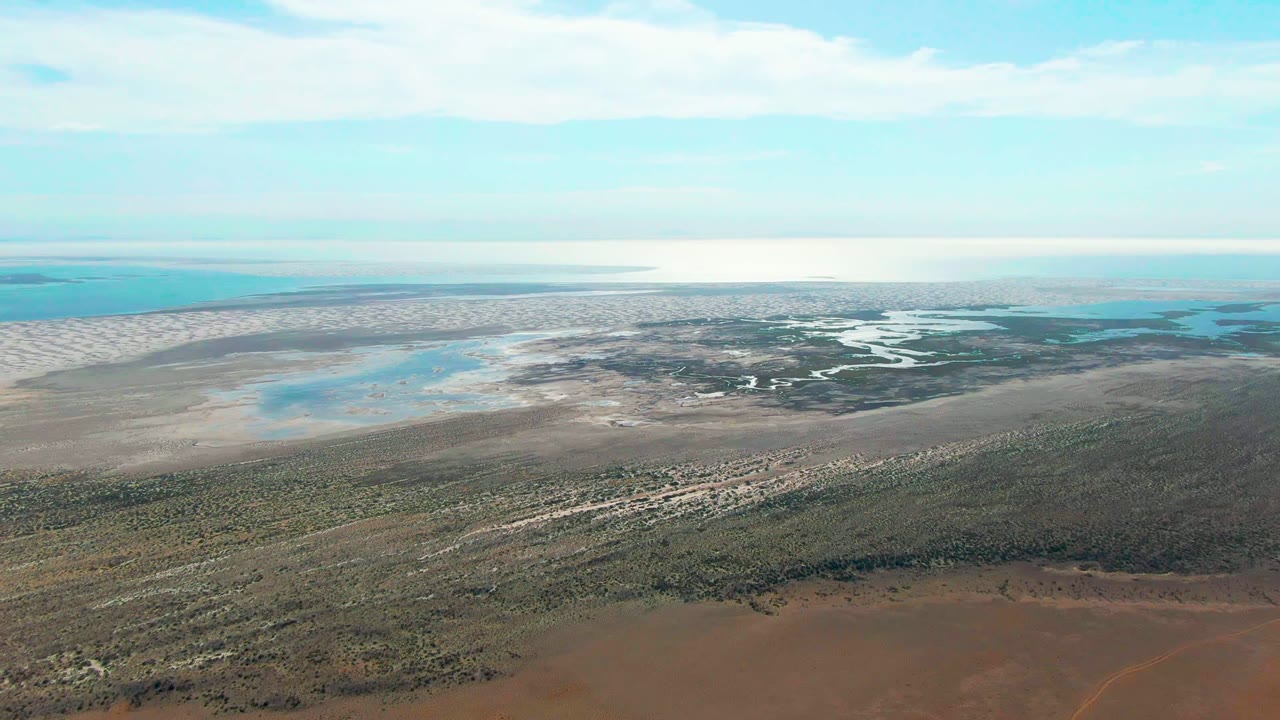 Wide aerial frame over Guerrero Negro wetlands with brown foreground scrub, pale sand flats, shallow pools, distant turquoise water, scattered clouds, and no visible people.