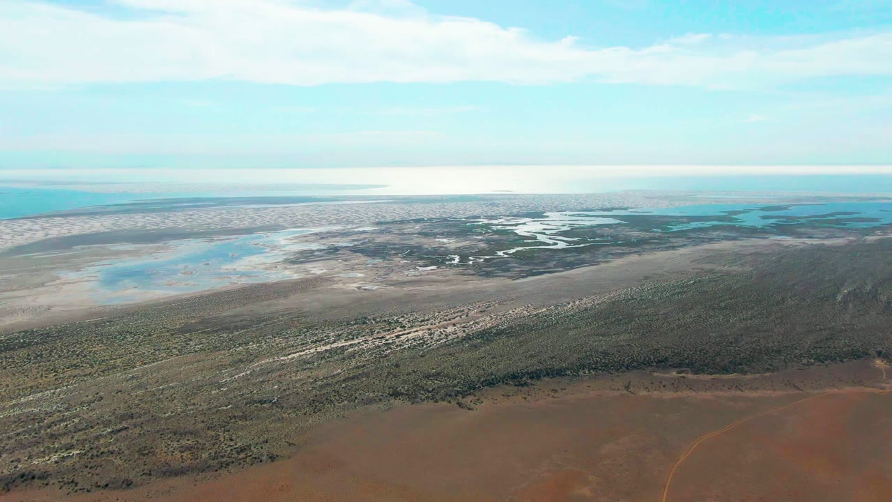 Wide aerial frame over Guerrero Negro coastal wetlands with red-brown flats in front, green scrub and pale sand beyond, blue water pools, ocean horizon, and no visible people.
