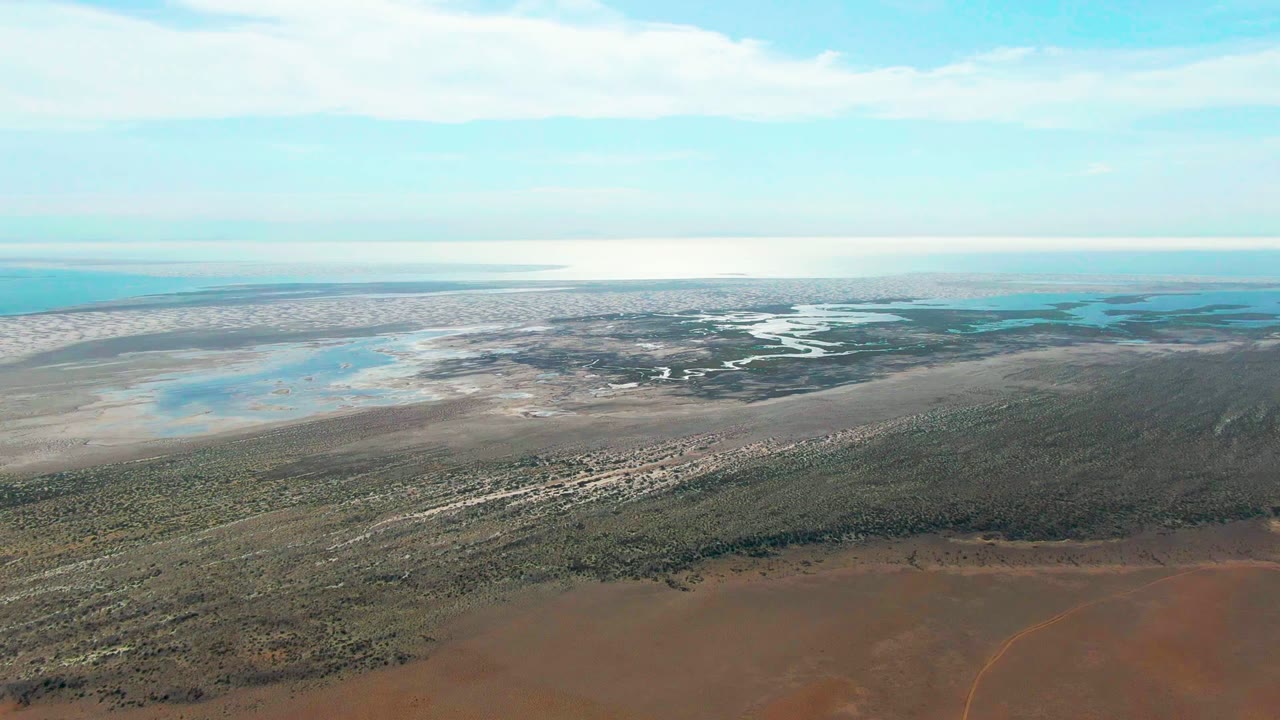 High aerial view across scrub, sandy wetland flats, and blue ocean water.