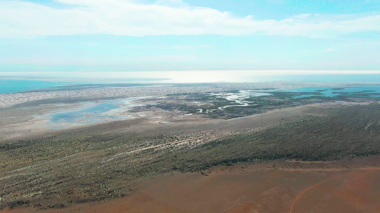 The aerial view over Guerrero Negro shows a red dirt foreground, textured green vegetation, pale sandbars around shallow water, distant ocean, clouded sky, and no visible people.