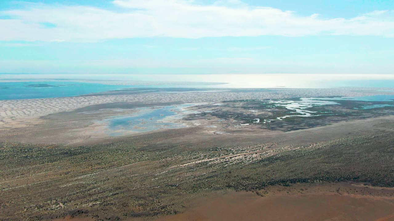 Aerial view over Guerrero Negro, Baja California Sur, showing scrubby foreground, exposed muddy flats, turquoise lagoon water, pale sandbars, and no visible people at the opening moment.