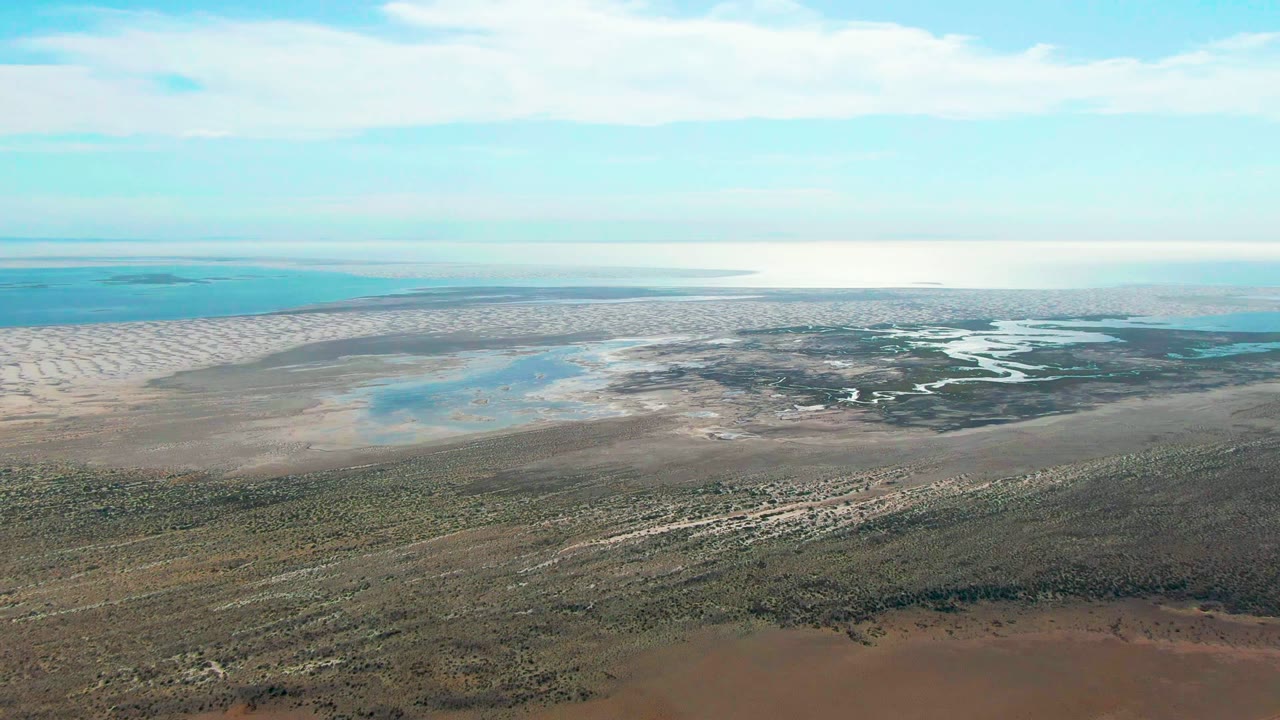Wide aerial lagoon view with muddy shoreline, pale sandbar, shallow water, and clouded blue sky.