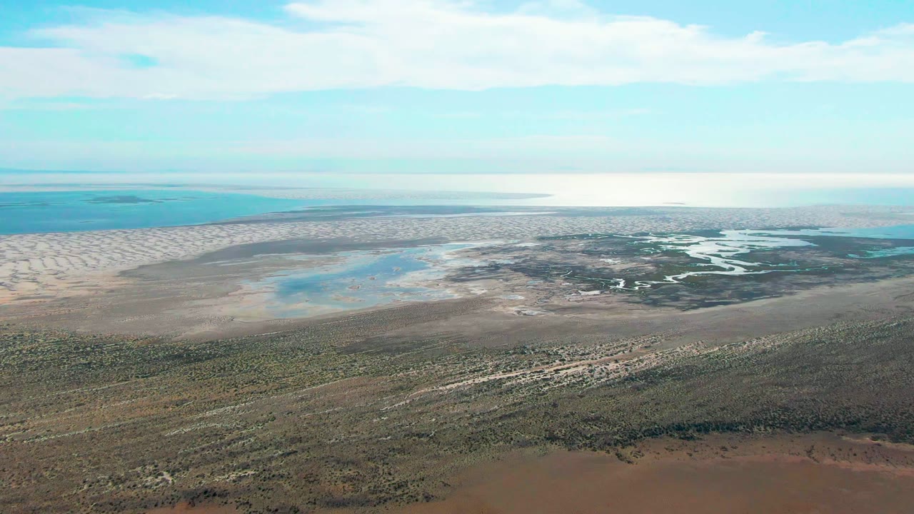 At 0:02, the Guerrero Negro lagoon remains in a high wide view, with shallow channels and darker wetland patches visible beyond the exposed muddy shore and no visible people.