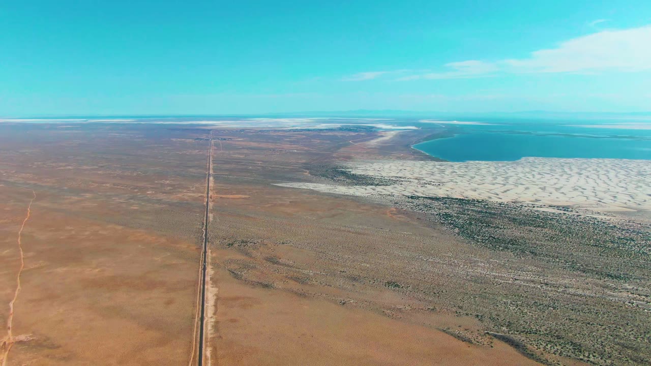 Elevated view over Guerrero Negro desert with a straight road leading toward the horizon, white salt lake edge to the right, turquoise water beyond it, and no visible people.