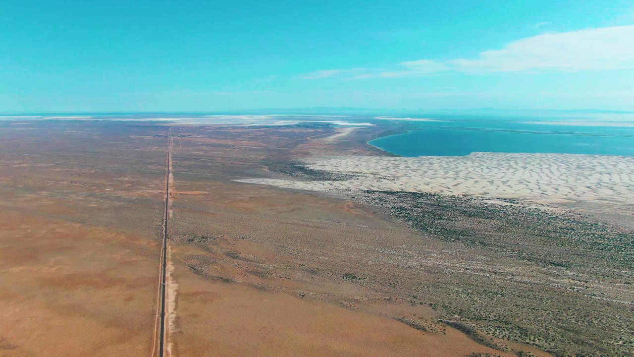 Aerial frame over Guerrero Negro, Baja California Sur, with a straight road crossing sandy desert toward white salt flats and turquoise water, no visible people.