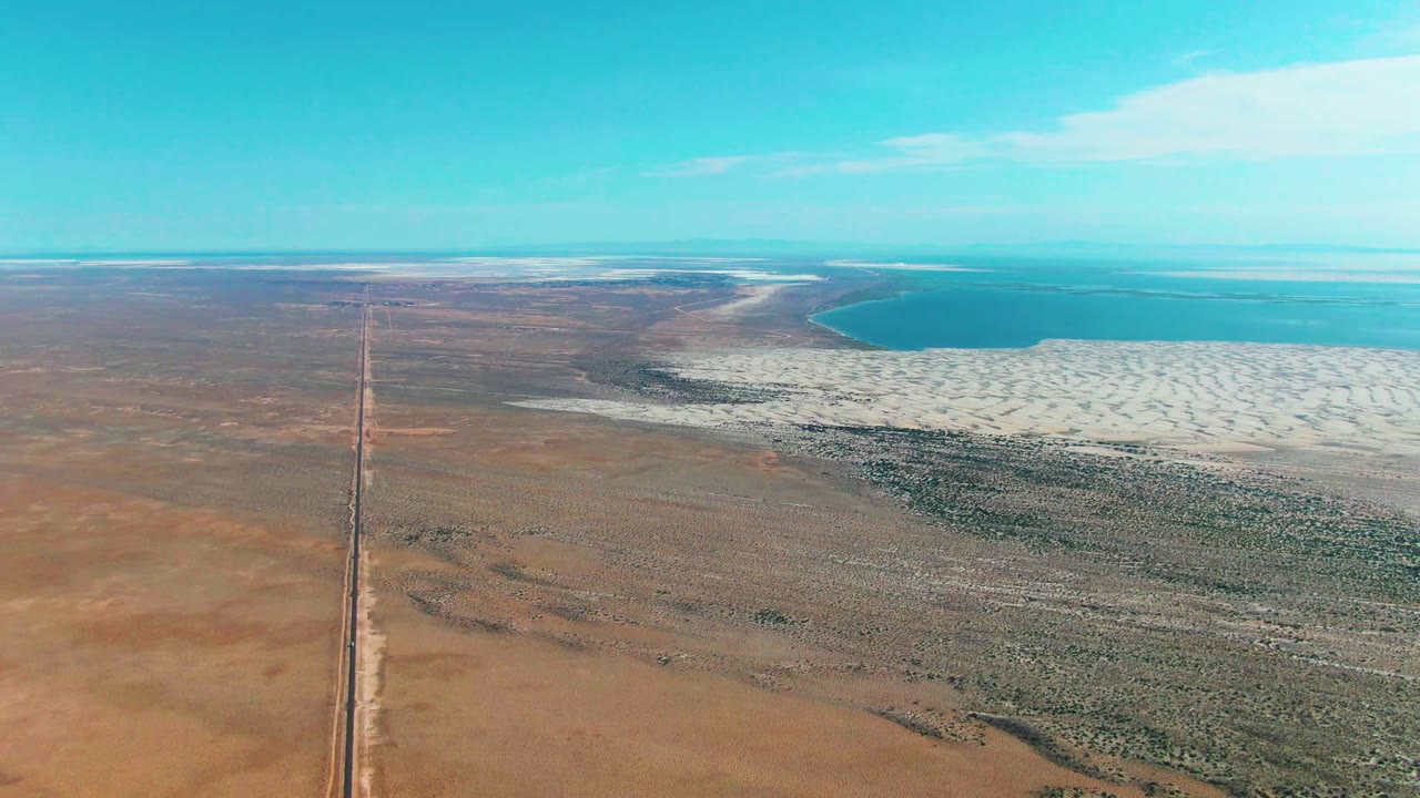 Straight road through arid terrain with salt flats and blue water ahead